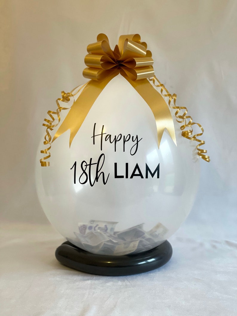 May include: A clear balloon with a gold ribbon and bow, filled with cash. The balloon has the text "Happy 18th LIAM" printed on it.