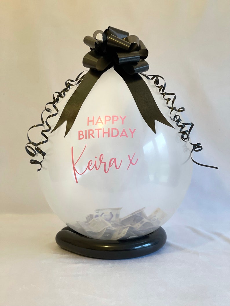 May include: A clear balloon with a black ribbon and bow, filled with cash. The balloon has the text "Happy Birthday Keira x" printed on it.