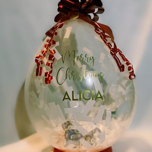 Puede incluir: Un globo transparente lleno de confeti blanco y dinero en efectivo, atado con una cinta roja y un lazo. El globo tiene una inscripción dorada que dice "Feliz Navidad Alicia".