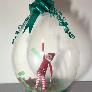 Puede incluir: Un globo de pl&aacute;stico transparente con un lazo y una cinta verdes, que contiene una mu&ntilde;eca de elfo con rayas rojas y blancas y un gorro verde. El globo est&aacute; lleno de confeti verde y rojo.