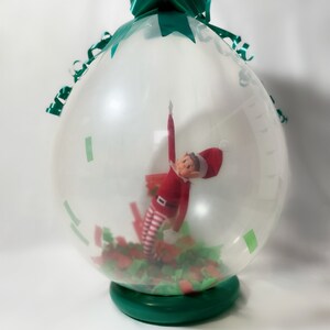 Puede incluir: Un globo transparente con un lazo verde en la parte superior, lleno de confeti verde y rojo y una peque&ntilde;a mu&ntilde;eca de elfo en su interior.