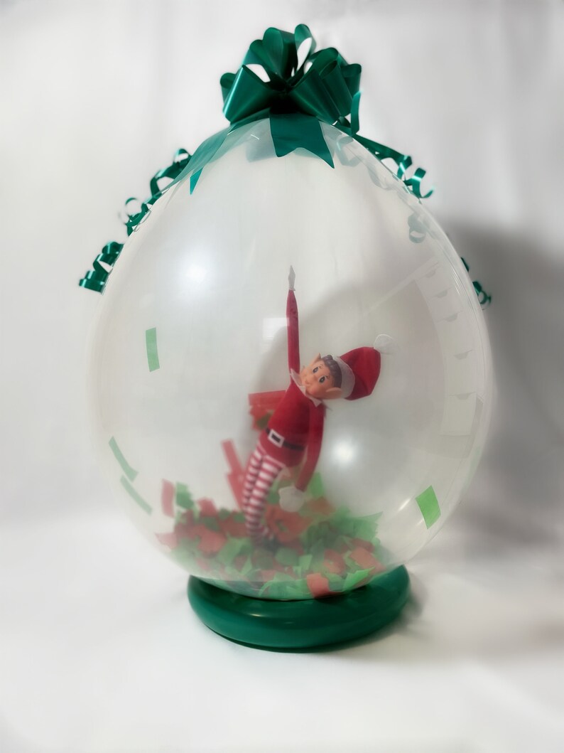 Puede incluir: Un globo transparente con un lazo verde en la parte superior, lleno de confeti verde y rojo y una peque&ntilde;a mu&ntilde;eca de elfo en su interior.