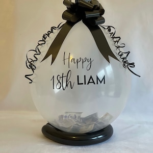 May include: A clear balloon with a black ribbon and bow, filled with cash. The balloon has the text "Happy 18th Liam" printed on it.
