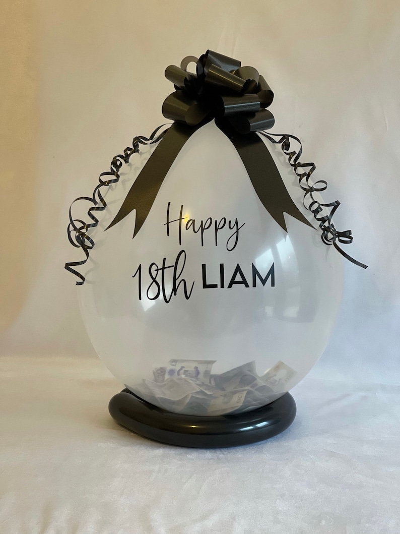 May include: A clear balloon with a black ribbon and bow, filled with cash. The balloon has the text "Happy 18th Liam" printed on it.