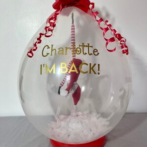 Puede incluir: Un globo transparente con un lazo rojo y una peque&ntilde;a mu&ntilde;eca de duende en su interior. El globo tiene una inscripci&oacute;n dorada que dice "Charlotte I'M BACK!"