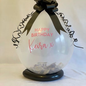 May include: A clear balloon with a black ribbon and bow, filled with cash. The balloon has the text "Happy Birthday Keira x" printed on it.