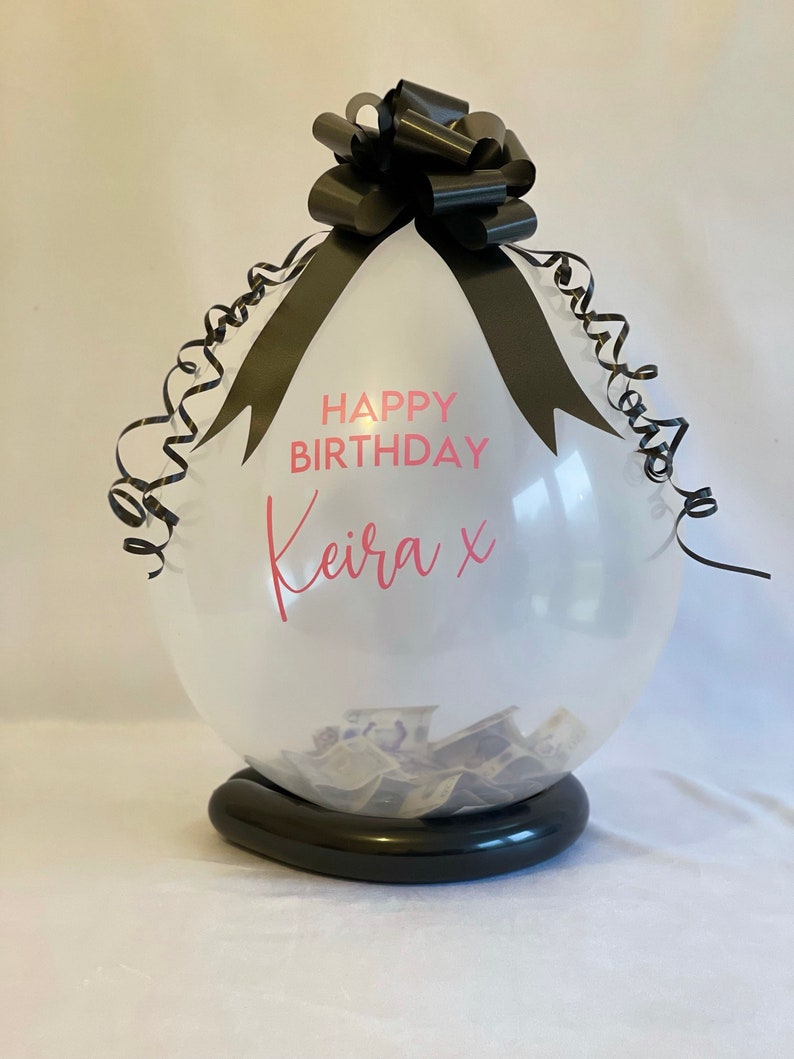 May include: A clear balloon with a black ribbon and bow, filled with cash. The balloon has the text "Happy Birthday Keira x" printed on it.
