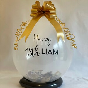 May include: A clear balloon with a gold ribbon and bow, filled with cash. The balloon has the text "Happy 18th LIAM" printed on it.