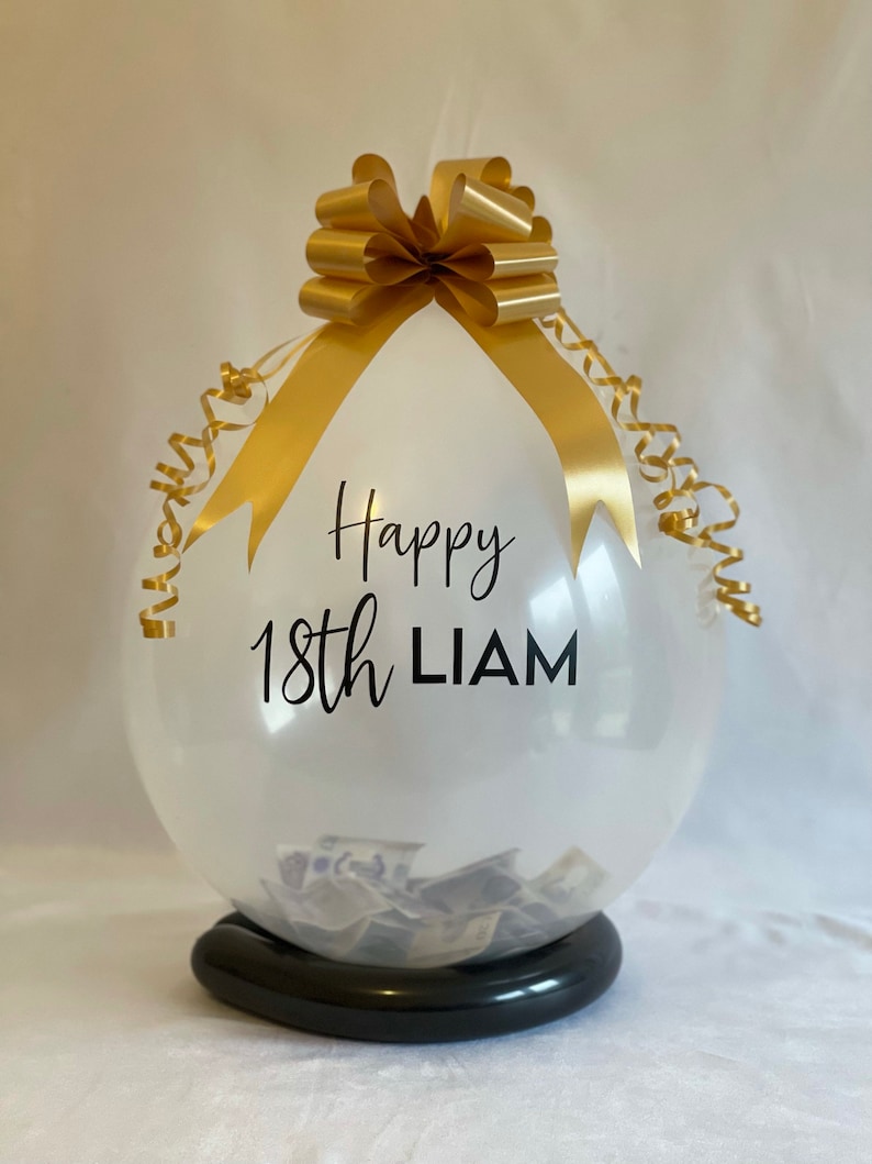 May include: A clear balloon with a gold ribbon and bow, filled with cash. The balloon has the text "Happy 18th LIAM" printed on it.