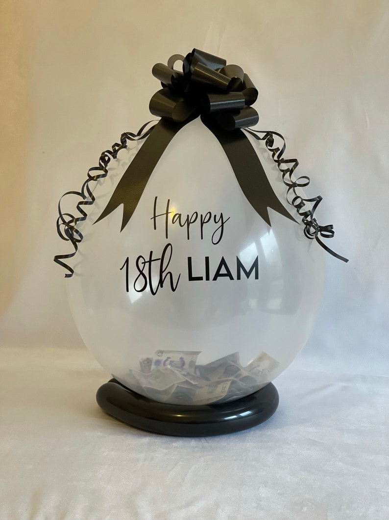 May include: A clear balloon with a black ribbon and bow, filled with cash. The balloon has the text "Happy 18th LIAM" printed on it.
