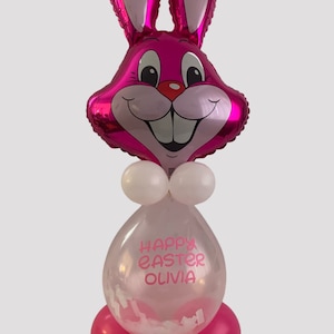 Peut inclure: Ballon en forme de lapin rose avec des oreilles blanches et un ballon transparent avec le texte "Joyeuses Pâques Olivia" et des confettis blancs à l'intérieur. Le ballon en forme de lapin est attaché au ballon transparent avec des ballons roses à la base.