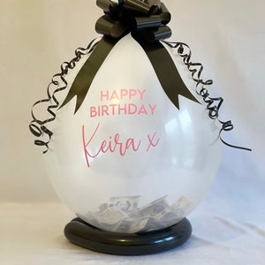 May include: A clear balloon with a black ribbon and bow, filled with cash. The balloon has the text "Happy Birthday Keira x" printed on it.