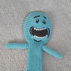 May include: A crocheted, light blue character with a large open mouth, white teeth, and black eyes. The character has outstretched arms and a long, cylindrical body. The item is handmade.