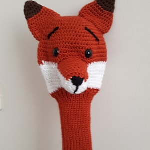 May include: A crocheted fox head golf club cover in orange, white, and brown. The fox has black eyes and a black nose.