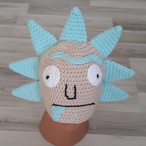 May include: A crocheted hat shaped like Rick Sanchez from the animated series Rick and Morty. The hat is beige with blue spikes and features a white face with black eyes and a black mouth.