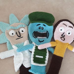 K&ouml;nnte beinhalten: Drei handgefertigte H&auml;kelpuppen, die jeweils eine Figur aus einer beliebten Zeichentrickserie darstellen. Die Puppen weisen unterschiedliche Gesichtsausdr&uuml;cke und Kleidungsdetails auf, darunter ein wei&szlig;er Laborkittel, eine gr&uuml;ne M&uuml;tze und ein gelbes Hemd. Die Puppen sind aus verschiedenen Garnfarben gefertigt.