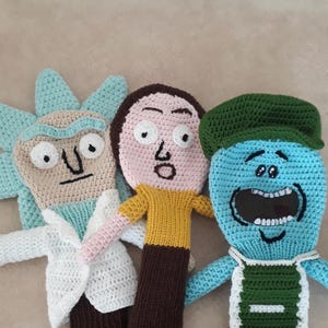 K&ouml;nnte beinhalten: Drei handgefertigte H&auml;kelpuppen, inspiriert von der Zeichentrickserie. Die Puppen weisen unterschiedliche Charakterdesigns auf: eine Figur mit hellblauen Haaren in einem wei&szlig;en Laborkittel, eine Figur mit braunen Haaren und einem gelben Hemd und eine blaue Figur mit einem gr&uuml;nen Hut.