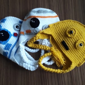 Puede incluir: Tres gorros de ganchillo, uno blanco con detalles azules y naranjas, uno blanco con detalles naranjas y grises, y uno amarillo con detalles negros.
