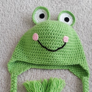 May include: Green crocheted frog hat with black eyes, pink cheeks, and a black stitched smile. The hat has long green crocheted ties.