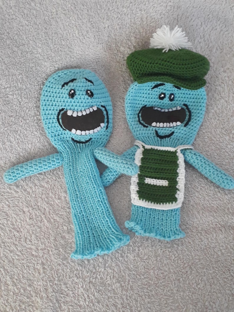 K&ouml;nnte beinhalten: Zwei hellblaue, geh&auml;kelte Charakterpuppen mit gro&szlig;en offenen M&uuml;ndern und schwarzen Schnurrb&auml;rten. Eine Puppe tr&auml;gt einen gr&uuml;nen Hut mit einem wei&szlig;en Bommel und eine gr&uuml;n-wei&szlig;e Weste. Die Puppen haben kleine Arme und werden auf einer grauen Oberfl&auml;che pr&auml;sentiert.