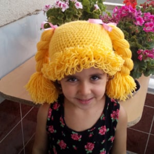 Può includere: Una persona giovane indossa un cappello all'uncinetto giallo con trecce. Il cappello ha un bordo arricciato e pompon ai lati delle trecce.
