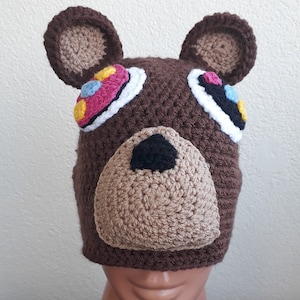 Puede incluir: Sombrero de oso de punto marrón con ojos coloridos y una nariz marrón. El sombrero está en una cabeza de maniquí.