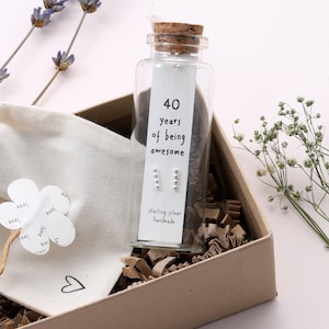 May include: A gift box containing a small glass bottle with sterling silver earrings. The bottle has a label that reads "40 years of being awesome". The box is filled with brown packing paper.