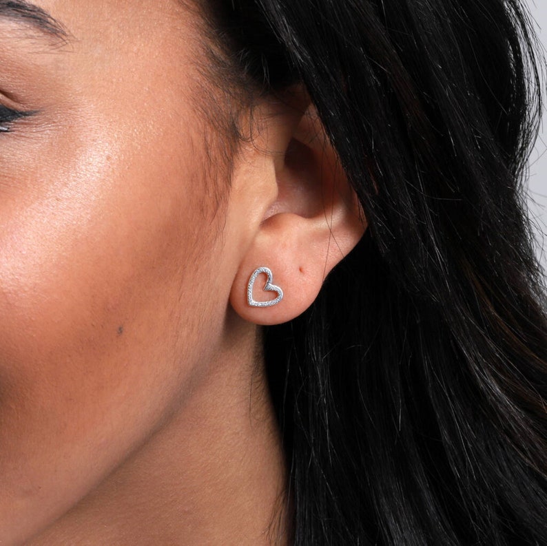May include: A silver-toned heart-shaped stud earring featuring a delicate outline design. The surface of the heart is textured with small, reflective metallic details that catch the light, creating a subtle sparkle against the earlobe.