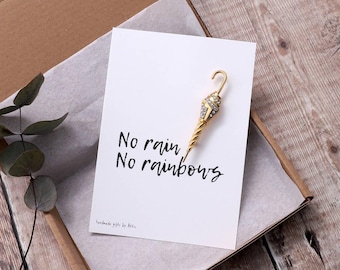 May include: A gold-colored umbrella-shaped brooch with crystal accents rests on a white card inside a cardboard box. The card reads "No rain, No rainbows" in black script. The box contains tissue paper and a sprig of greenery.