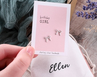 May include: A pink card with the text "birthday GIRL" and "sterling silver handmade" on it. Two silver bow earrings are displayed on the card. The card is sitting on a white drawstring bag with the name "Ellen" written on it.