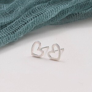 May include: A pair of silver-toned stud earrings shaped like open hearts. Each earring features a textured, granular surface finish on the metal frame and includes a standard post backing for secure attachment to the earlobe.