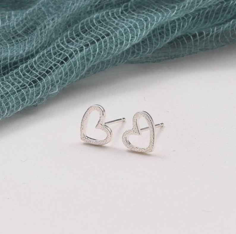 May include: A pair of silver-toned stud earrings shaped like open hearts. Each earring features a textured, granular surface finish on the metal frame and includes a standard post backing for secure attachment to the earlobe.