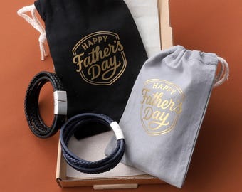 May include: A Father's Day gift set featuring two leather bracelets in black and navy blue, with silver clasps. The bracelets are presented in a cardboard box with two drawstring bags, one black and one gray, each printed with "Happy Father's Day" in gold.