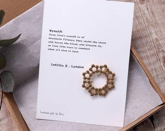 May include: A gold-colored wreath made of small flower shapes. The wreath is on a white card with a poem about true love's wreath. The poem reads: "True love's wreath is of mountain flowers, they stand the storm and brave the blast, and blossom on, so love like ours is sweetest when all else is past." The poem is signed "Letitia E. Landon".
