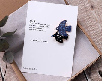 May include: A blue and black enamel pin featuring two birds, one perched on top of the other. The pin is accompanied by a card with a poem by Alexander Posey titled "Bird".