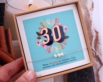 May include: A small gift box with a blue background and the number 30 in large, colorful letters. The box contains a silver chain necklace with three white pearls. The text on the box reads "Three PEARLS to celebrate three precious and distinguished decades".