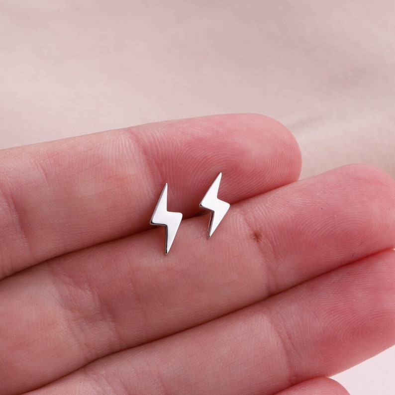 Puede incluir: Un par de peque&ntilde;os pendientes de bot&oacute;n con forma de rayo en tono plateado pulido. Cada pendiente presenta un dise&ntilde;o angular y n&iacute;tido con una superficie met&aacute;lica lisa y reflectante, presentados como un conjunto de dos piezas individuales.