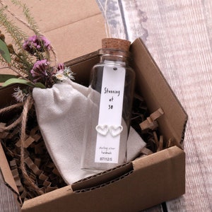 Puede incluir: Una peque&ntilde;a botella de vidrio con un tap&oacute;n de corcho contiene un par de pendientes de plata en forma de coraz&oacute;n. La botella tiene una etiqueta blanca con el texto "Stunning at 30", "sterling silver handmade" y "BT112/S". La botella est&aacute; sentada en una caja de cart&oacute;n marr&oacute;n con papel triturado.