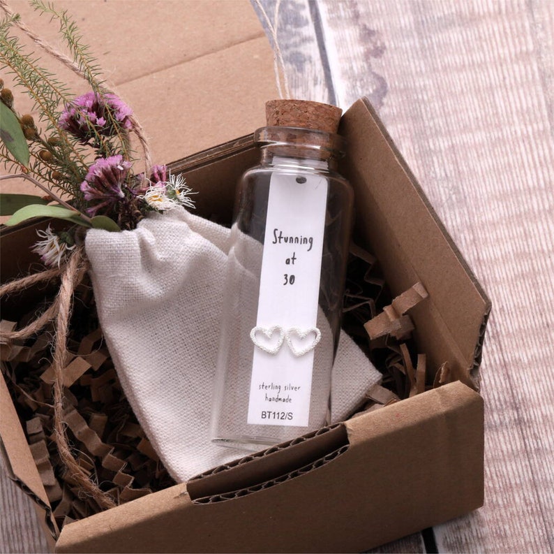Puede incluir: Una peque&ntilde;a botella de vidrio con un tap&oacute;n de corcho contiene un par de pendientes de plata en forma de coraz&oacute;n. La botella tiene una etiqueta blanca con el texto "Stunning at 30", "sterling silver handmade" y "BT112/S". La botella est&aacute; sentada en una caja de cart&oacute;n marr&oacute;n con papel triturado.