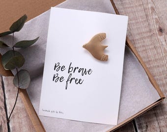 May include: A white card with the text "Be brave Be free" and a gold bird figurine on top. The card is in a brown box with white tissue paper.