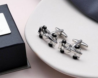 May include: A pair of silver-toned cufflinks shaped like race cars, with black wheels, displayed on a white plate. A black box with a silver label is in the background.