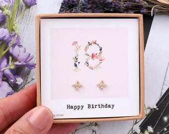 May include: A small gift box with a pink background and the number 18 in floral design. The box contains a pair of small gold earrings with pink stones. The text "Happy Birthday" is printed on the box.