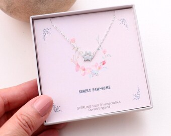 May include: A silver paw print pendant necklace displayed in a square gift box. The box features a light pink background with a floral wreath illustration, the text SIMPLY PAW-SOME, and the words STERLING SILVER hand crafted Dorset England.