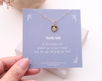 May include: A gold-toned heart-shaped St. Christopher pendant on a delicate chain, displayed on a light purple card with the text TRAVEL SAFE, St Christopher will protect you on your travels keep you safe and guide you home, STERLING SILVER hand crafted Dorset England.