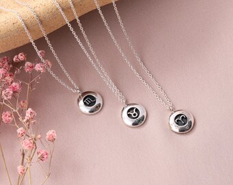 May include: Silver necklaces with round pendants featuring zodiac symbols. The pendants have a black background with the zodiac symbol in silver. The necklaces are displayed on a pink surface with pink flowers.