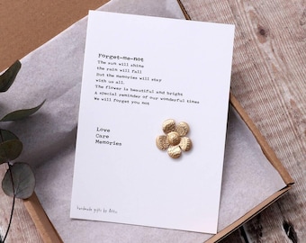 May include: A white card with a poem that reads "Forget-me-not The sun will shine the rain will fall But the memories will stay with us all. The flower is beautiful and bright. A special reminder of our wonderful times We will forget you not Love Care Memories" and a gold flower charm.