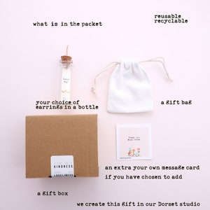 Puede incluir: Una caja marr&oacute;n con las palabras "A Little Bit of Kindness" y "Packed with Loveliness" en ella. Una peque&ntilde;a botella de vidrio con un tap&oacute;n de corcho contiene aretes. Tambi&eacute;n se incluye una bolsa de cord&oacute;n blanco, una tarjeta de agradecimiento y una caja de regalo. El texto "reusable recyclable" est&aacute; en la parte superior de la imagen.