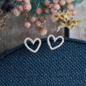 May include: A pair of silver-toned heart-shaped stud earrings with a textured, granular surface finish. The open-frame heart design features a slightly irregular, hand-crafted appearance, showcasing a metallic sheen against a dark blue fabric.
