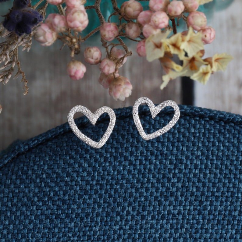 May include: A pair of silver-toned heart-shaped stud earrings with a textured, granular surface finish. The open-frame heart design features a slightly irregular, hand-crafted appearance, showcasing a metallic sheen against a dark blue fabric.