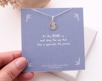 Puede incluir: Un collar de plata de ley con un pequeño colgante circular con un diseño en relieve, presentado en una tarjeta cuadrada azul con el texto: It's the FRIENDS we meet along the way that help us appreciate the journey. STERLING SILVER hand crafted Dorset England.